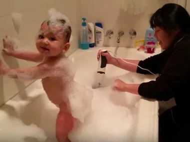 A child enjoys a bubble bath while a woman uses electric beaters to create more bubbles in the tub.