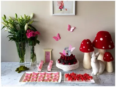 Fairy-theme birthday setup with cake, butterfly decor, mushrooms, bunny statue, and flowers on a marble table.