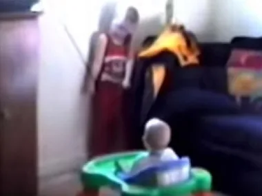A toddler stands near a blind cord, with a baby in a walker watching, highlighting a potential choking hazard.