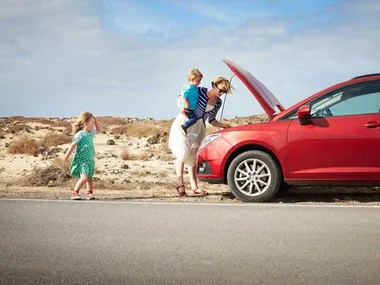 A woman with two children checks under the hood of a red car on a desert road.