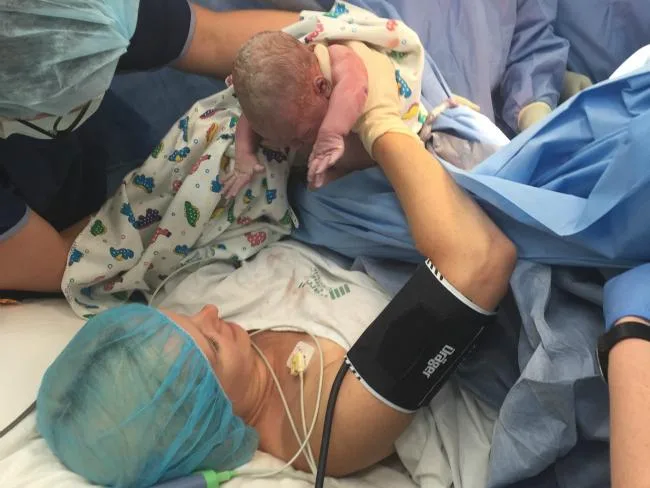 A mother assists in delivering her newborn during a maternal-assisted caesarean, surrounded by medical staff.