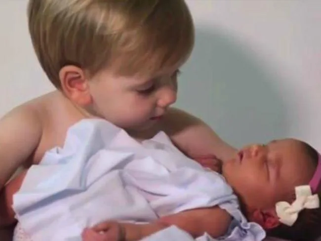Toddler boy gently holding his newborn baby sister, both wrapped in white cloth, with loving expressions.