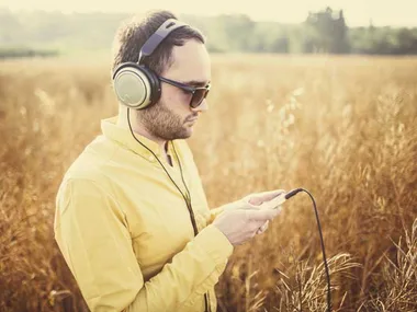 Man in yellow shirt wearing headphones and sunglasses, standing in a field, using a mobile device.