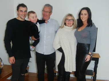 A family of five posing indoors, including a toddler held by a man, with two women and another man, smiling at the camera.