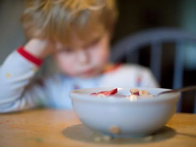 A young child gazes sadly at a bowl of cereal with strawberries at a table.