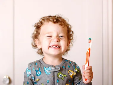 Toddler with curly hair winking, holding a toothbrush, wearing pajamas with guitar prints, smiling broadly.