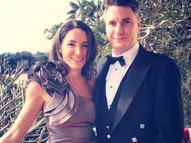 Two people smiling, with the woman in a ruffled dress and the man in a tuxedo, standing outdoors with trees in the background.