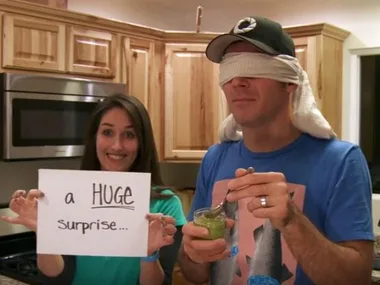 Woman holds "a HUGE surprise" sign; man blindfolded with spoon in kitchen.