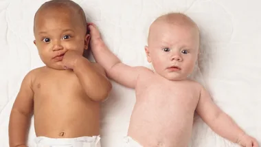 Two babies lying on a white surface, one with darker skin, sucking fingers, and one with lighter skin, looking forward.