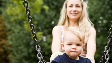 Toddler on a swing with a smiling woman in the background, outdoors on a sunny day.