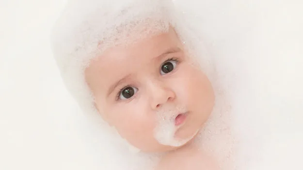 Baby with wide eyes covered in white fluffy bubbles, floating in a bath.