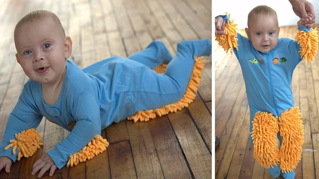 Baby wearing a blue onesie with orange mop-like fringes, crawling and standing on a wooden floor.