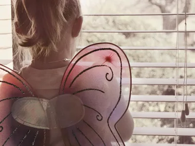 Young child wearing butterfly wings looks out a window with blinds to an outdoor view.