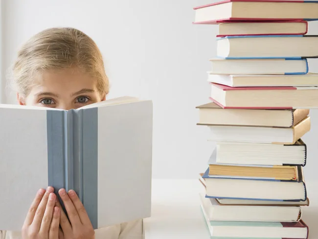 Girl peeks over open book with stack of books beside her on table.