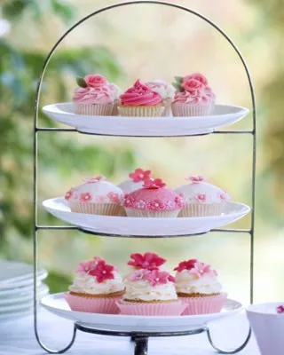 Three-tier stand with pink and white frosted cupcakes, decorated with flowers and leaves, outdoors.
