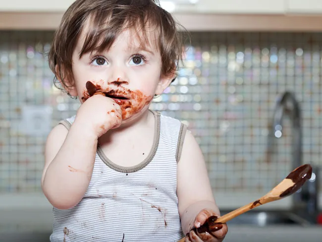 Toddler in a striped tank top with chocolate smeared on face, holding a chocolate-covered wooden spoon in a kitchen.