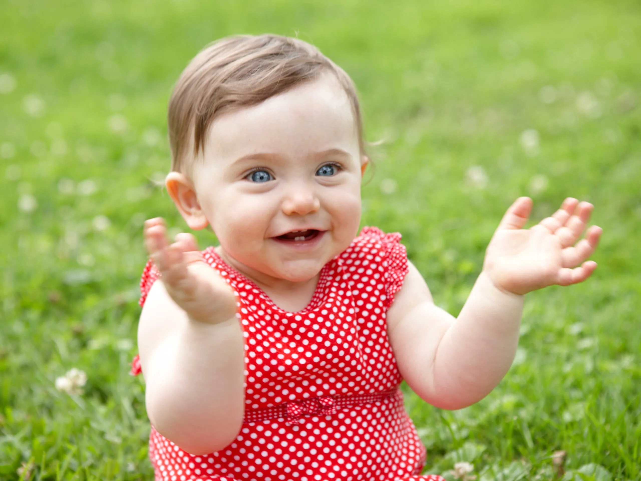 A smiling baby sitting on grass, wearing a red polka dot dress, with hands raised in a playful gesture.