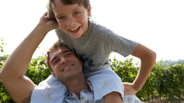 Father carrying smiling child on shoulders in a sunny vineyard.