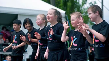 Children with Down syndrome joyfully dancing in matching black shirts with colorful logos, outdoors under a tent.