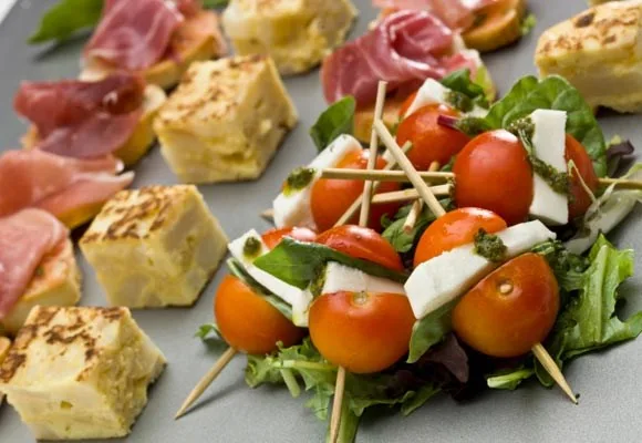 Assorted tapas, including cherry tomato skewers with cheese and mixed greens, set on a gray platter.