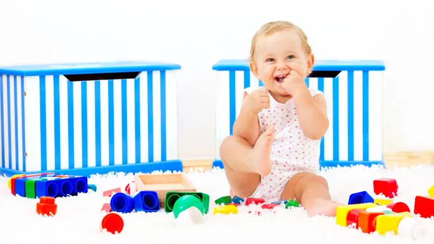 Baby smiling and sitting on a white carpet, surrounded by colorful toy blocks with blue-striped storage boxes in the background.