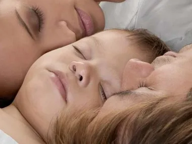 Three people peacefully asleep in bed, including a child between two adults, close-up view of their faces.