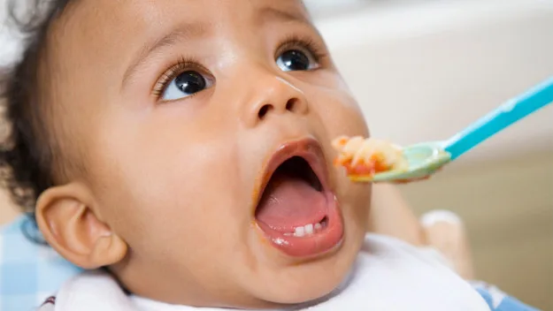 A baby with an open mouth being fed with a spoonful of puree or baby food.