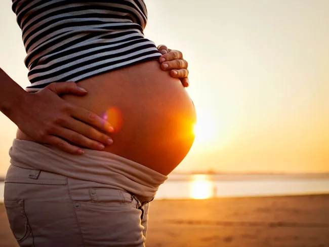 Pregnant woman holds her belly on a beach at sunset, with the sunlight creating a warm glow.