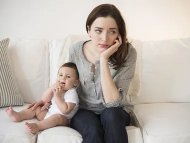 A mother looks worried while sitting on the couch with her baby.