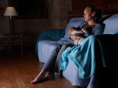 Mother sitting on a sofa in a dimly lit room, feeding a baby wrapped in a blanket.