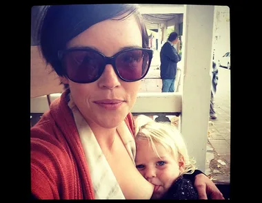 A woman wearing sunglasses breastfeeds a young child while sitting outdoors.