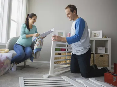 Pregnant woman and man assemble a crib in a nursery, smiling and checking instructions, surrounded by baby items.