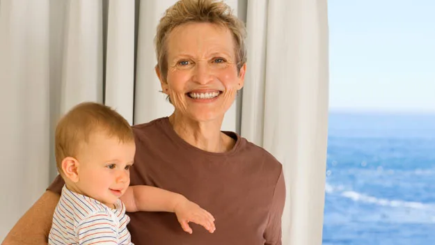 Older woman smiling, holding a baby by the sea with a curtain behind them.