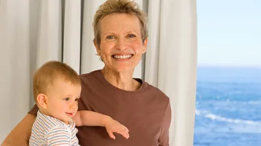 Older woman smiling, holding a baby by the sea with a curtain behind them.