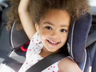 Girl smiling in a car seat, secured with a seatbelt; curly hair and white polka dot dress.
