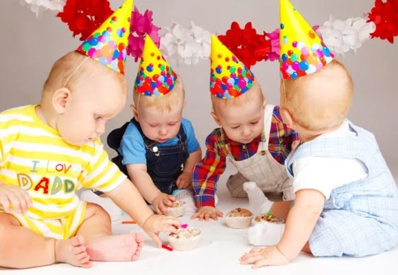 Babies at a birthday party wearing colorful hats, playing with cupcakes on the floor, with festive decorations in the background.