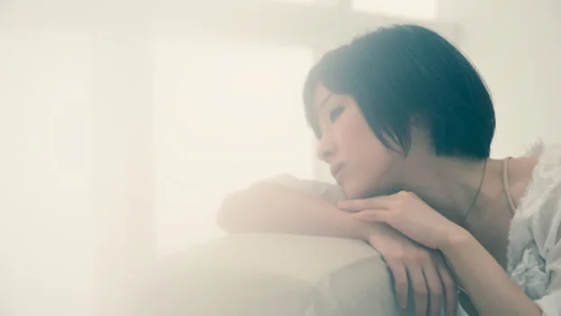 A woman with short hair leans on a couch, gazing thoughtfully through a bright, soft-focus background.