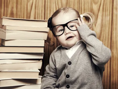 Toddler with glasses and sweater sitting beside a stack of books, smiling and touching glasses.