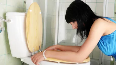 Woman in blue dress leaning over a toilet, appearing to be nauseous in a tiled bathroom.