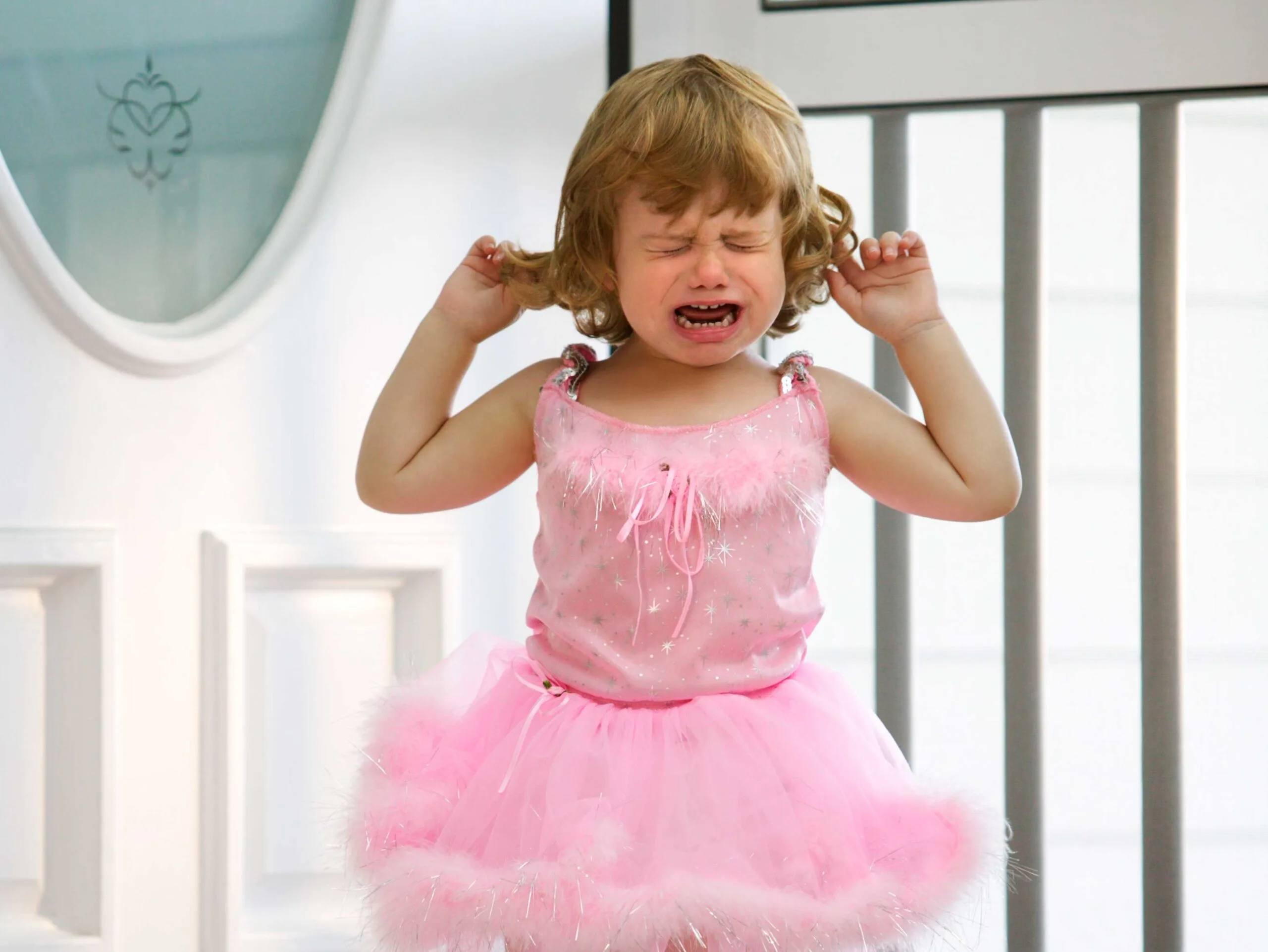 A toddler wearing a pink tutu cries and holds her hair, standing near a white door.