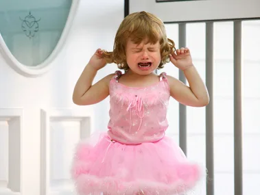 A toddler wearing a pink tutu cries and holds her hair, standing near a white door.