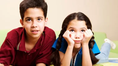 Two children indoors; a boy looks up while a bored girl with braided hair rests her chin on her hands.