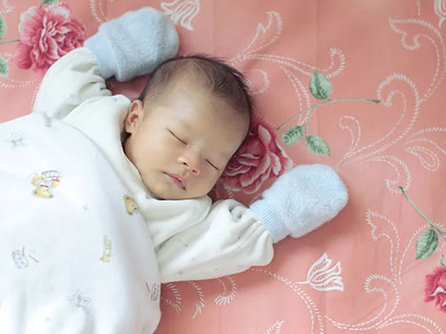 Sleeping baby in white outfit and blue mittens on a pink floral blanket.