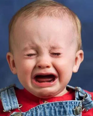 A crying baby wearing denim overalls and a red shirt against a blue background.