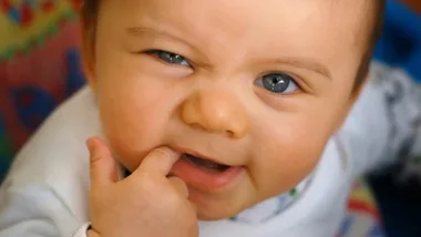 Baby with finger in mouth, appearing to teeth, wearing light-colored clothing on a colorful background.