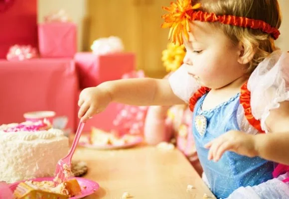 Toddler dressed as a princess, with an orange headband, eating cake with a pink fork at a birthday party.