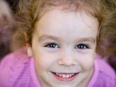 A young child with curly hair in a pink shirt smiling brightly at the camera.