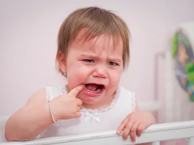 Crying toddler in a crib wearing a white dress, with a finger in their mouth, appearing upset.