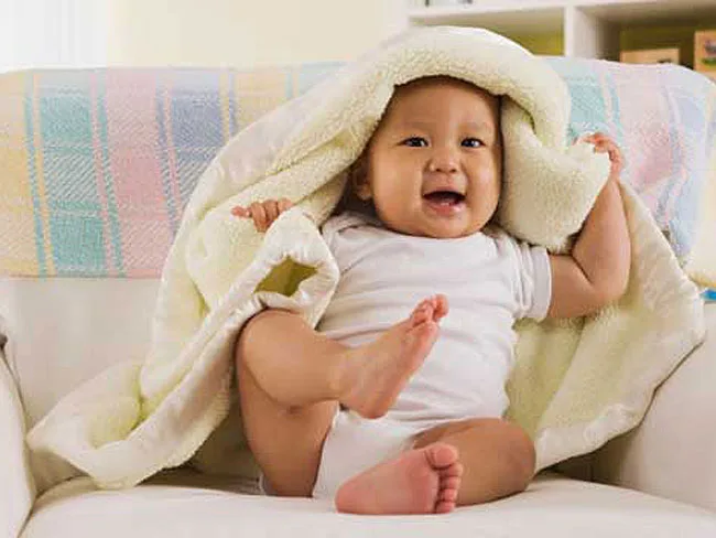 A happy baby playing under a soft blanket, wearing a white onesie, sitting on a couch.