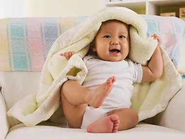 A happy baby playing under a soft blanket, wearing a white onesie, sitting on a couch.